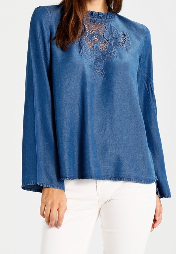 Blouse en jean bleu à manches longues, ornée d'une broderie florale délicate sur la poitrine et d'un col à volants. Texture lisse, coupe ample.