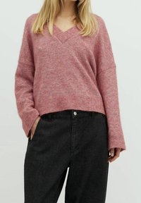 Rosa, strukturierter V-Ausschnitt-Pullover mit lockerer Passform und langen Ärmeln, kombiniert mit hochgeschnittenen, dunklen Denim-Hosen mit Knopfverschluss vorne.