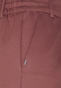 Primer plano del bolsillo de un pantalón de tela burdeos con cintura elástica y una pequeña etiqueta blanca "ONLY" cerca de la costura del bolsillo.