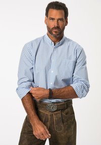 Camisa de cuadros azul y blanca, con las mangas remangadas, combinada con pantalones de cuero marrón que presentan un hebilla decorativa y detalles en los bolsillos.