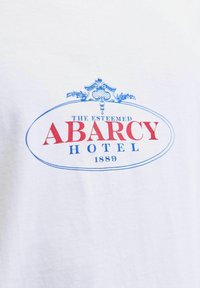 Fehér póló kék és piros grafikus dizájnnal. A szöveg olvashatóan így szól: "A MEGTISZTELT ABARCY HOTEL 1889" egy díszes ovális keretben.
