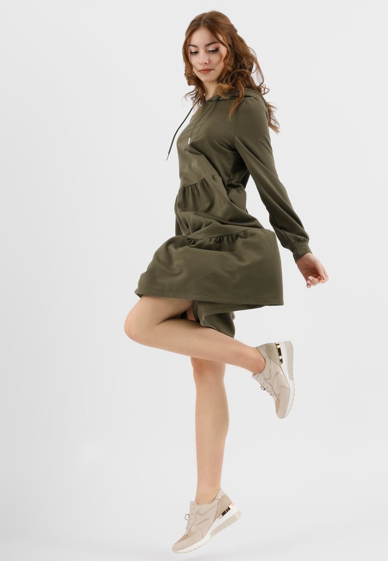 Abito verde oliva con cappuccio e orlo arricciato, abbinato a sneakers beige con dettaglio dorato. Tessuto morbido, maniche lunghe e vestibilità rilassata.