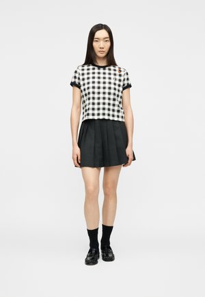 Jeune femme portant un haut à manches courtes à carreaux noir et blanc, une jupe plissée noire, des chaussettes noires et des mocassins noirs, debout devant un fond uni.