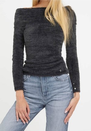 Femme portant un pull noir ajusté, à manches longues et épaules dénudées, en tissu duveteux, avec un jean taille haute bleu clair, les mains reposant sur les hanches.