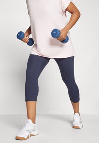 Lys rosa treningstopp, marineblå leggings, holder blå hantler. Hvite sko med grå detaljer. Fokuserer på materiale og passform for trening.