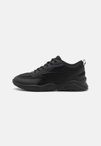 Black athletic shoes with a textured upper, padded collar, and a thick rubber sole. Features a minimalistic design and contrasting accents.