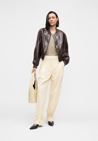 Brown shiny bomber jacket over a beige top, paired with high-waisted cream trousers and black pointed shoes. Holding a cream bag.