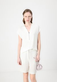 Hvit kortermet topp med dyp V-hals og glidelåsdetalj, sammen med lyse beige shorts og en myk rosa håndveske.
