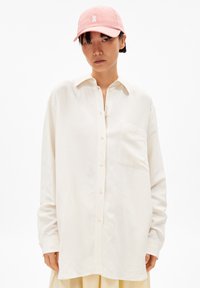 Weißes Langarmhemd mit geknöpftem Vorderteil und einer Brusttasche, kombiniert mit einer pastellrosa Kappe mit Logo. Flacher Hintergrund.