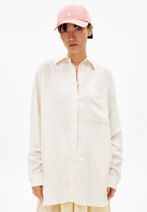 Witte lange mouw shirt met een knoopsluiting aan de voorkant en een borstzak, gecombineerd met een pastelroze pet met een logo. Effen achtergrond.