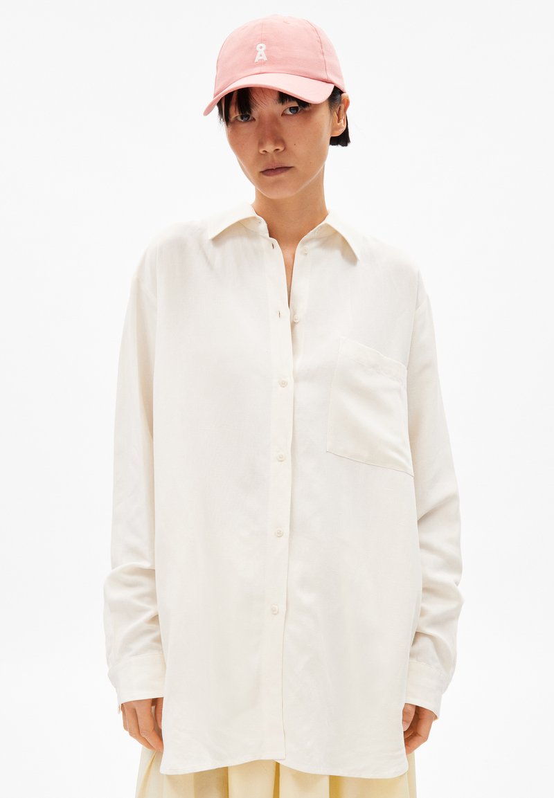 Weißes Langarmhemd mit geknöpftem Vorderteil und einer Brusttasche, kombiniert mit einer pastellrosa Kappe mit Logo. Flacher Hintergrund.