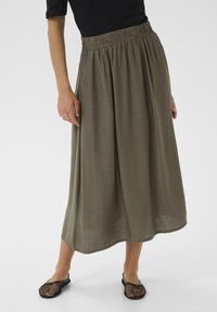 Jupe maxi en tissu vert olive léger et texturé, dotée d'une taille élastique et d'une silhouette ample et fluide. Portée avec des chaussures en mesh noires.