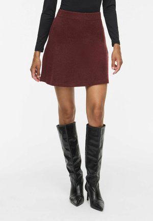 Femme portant une jupe trapèze rouge foncé avec un haut noir à manches longues et des bottes en cuir noires montantes aux genoux sur un fond uni.