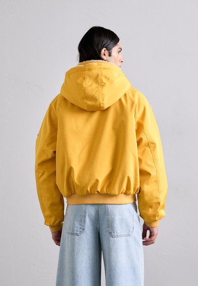 Yellow hooded jacket with a soft texture, elasticated cuffs and hem. Paired with light blue wide-leg jeans. Back view.