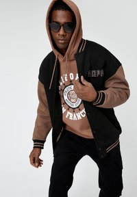 Chaqueta de béisbol negra con mangas marrones, llevada sobre un sudadero marrón con un diseño gráfico. Unas gafas de sol oscuras completan el look.