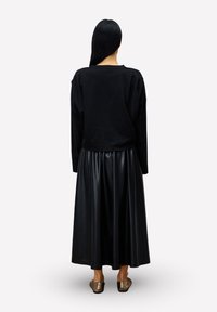 Frau mit langen schwarzen Haaren, die ein lockeres schwarzes Oberteil, einen glänzenden schwarzen Midirock und metallische spitze Ballerinas trägt und mit dem Rücken zur Kamera steht.