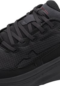 Baskets de sport noires en tissu mesh, semelle épaisse, lacets et détails subtils de la marque sur la languette et le côté.