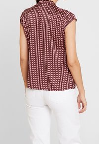 Frau in einem ärmellosen bordeauxfarbenen gemusterten Bluse, kombiniert mit weißen Hosen, von hinten vor einem schlichten Hintergrund betrachtet.