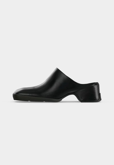 Mule en cuir noir avec un petit talon, un bout arrondi et un design sans fermeture, présentée en profil latéral sur un fond clair.