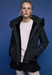 Črna puhovka z kapuco, krznenim robom, zadrgo in sprednjimi žepi, nošena preko svetlega puloverja in večplastne krila na modrem ozadju.