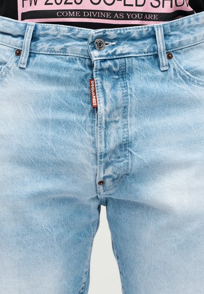 Jean en denim bleu clair avec un bouton en métal, passants de ceinture visibles et une étiquette rouge DSQUARED2 près de la fermeture éclair, porté avec une chemise rose et noire rentrée.