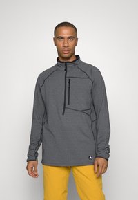 Maglione grigio con zip a quarto, caratterizzato da un tessuto texturizzato, una tasca sul petto e dettagli di cucitura a contrasto. Abbinato a pantaloni gialli.
