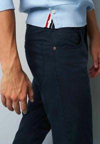 Marineblauwe katoenen broek met zijzakken en een koperen knoopdetail. De hand van het model rust op de stof, wat de gladde textuur benadrukt.