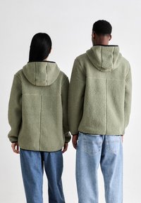 Twee personen in bijpassende olijfgroene fleece jassen met capuchons, voorzien van een gestructureerd oppervlak en verlaagde schouders, gecombineerd met blauwe spijkerbroeken.