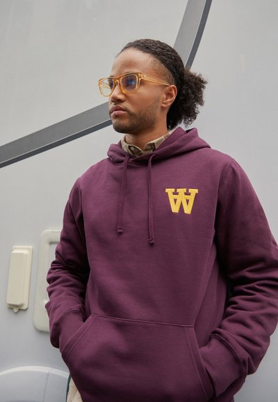 Körsbärsröd hoodie med en gul broderad "W"-logotyp, tillverkad av slät tyg. Har en framficka och dragsko över huvan. Klär i klara glasögon.