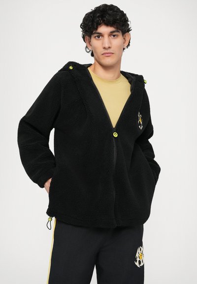 Fekete bolyhos dzseki cipzárral, logóval kiemelve. Kapucnival és elasztikus mandzsettákkal felszerelt, fekete nadrággal, mely sárga csíkokkal díszített.