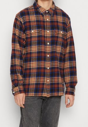 Personne portant une chemise à carreaux boutonnée à manches longues avec des couleurs marron, beige, bleu et rouge, associée à un jean gris foncé.