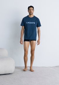 Tamno plava pamučna majica s "LACOSTE" logotipom, uparen s crnim kratkim hlačama. Stojeći na sivoj tepih, bosi, neutralna pozadina.