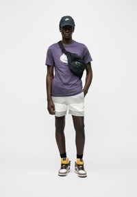 Άνδρας που φοράει μωβ μπλουζάκι Nike, γκρι και λευκά σορτς, μαύρο καπέλο Nike, μαύρη τσάντα χιαστί και πολύχρωμα sneakers ψηλού πάτου, στέκεται μπροστά σε λευκό φόντο.