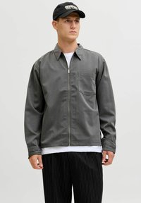 Chaqueta gris con cremallera y bolsillo en el pecho, con cuello con solapas y mangas largas. Combinada con pantalones negros de rayas finas y una gorra negra.