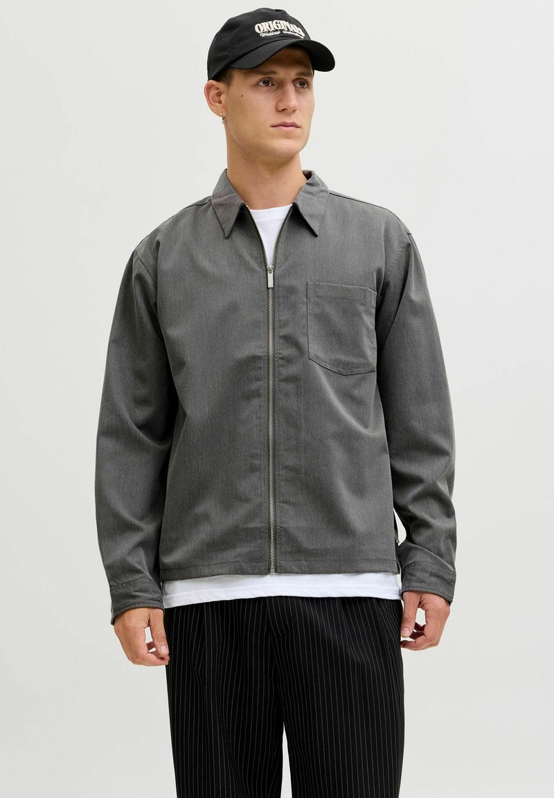 Chaqueta gris con cremallera y bolsillo en el pecho, con cuello con solapas y mangas largas. Combinada con pantalones negros de rayas finas y una gorra negra.