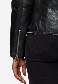 Zwarte leren jas met een cropped ontwerp, ritsmanchetten en ruwe randdetails. Draag over donkerdenim broek met een achterzak.