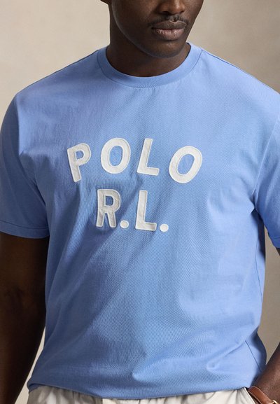 Άνδρας που φοράει ανοιχτό μπλε T-shirt με λευκά κεντημένα γράμματα "POLO R.L." στο στήθος, συνδυασμένο με ανοιχτόχρωμο παντελόνι.
