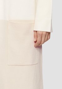 Helles, cremfarbiges Strickkleid mit strukturierter Oberfläche, langen Ärmeln und einer großen Tasche im unteren Bereich. Weiches, minimalistisches Design.