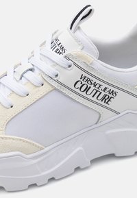 Sneaker bianco che presenta un mix di materiali: tessuto liscio, suede e suola in gomma. Include un logo su un'etichetta in pelle e lacci robusti.
