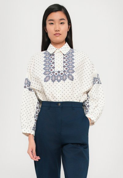 Blusa branca com bordados florais azuis e pequenos padrões, complementada por calças escuras de cintura alta. Gola com botões, mangas longas.