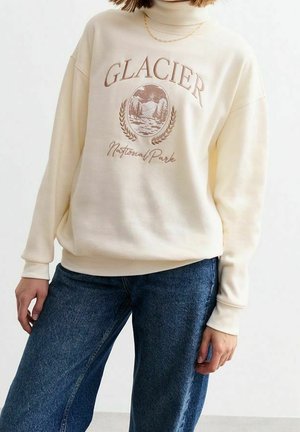 Person iført en cremefarvet højhalset sweatshirt med teksten "Glacier National Park" og bjerggrafik, kombineret med blå jeans og en guldkæde.