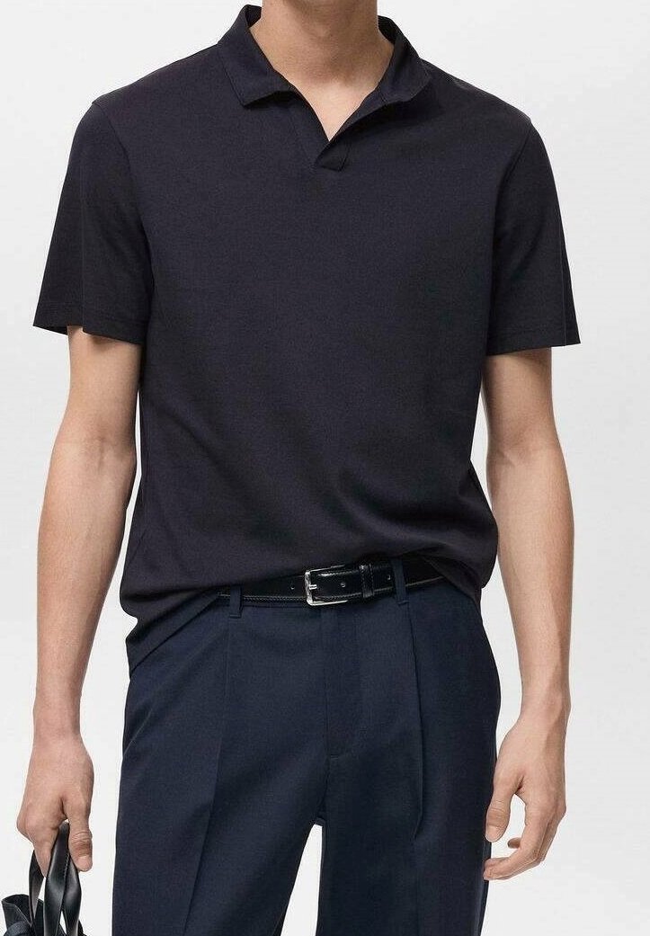 Homme portant un polo noir cintré à manches courtes, rentré dans un pantalon slim foncé avec une ceinture noire, tenant des sandales noires.