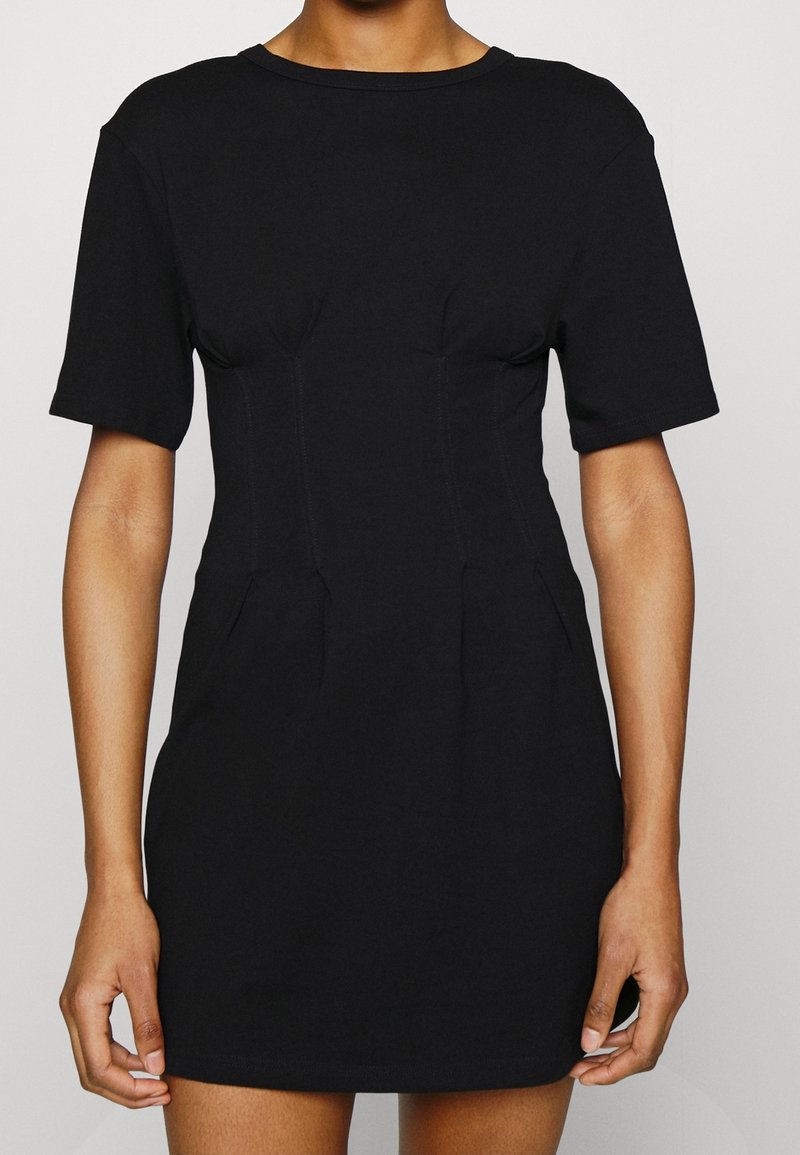 Personne portant une robe noire ajustée à manches courtes avec des détails de couture et un col rond, debout devant un fond clair.