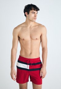 Rote Badehose mit einem Kordelzugbund, ausgestattet mit navyblauen und weißen horizontalen Streifen auf der Vorderseite. Leichter Stoff, bis über das Knie.