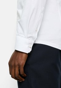 Weißes Baumwollhemd mit Knopfmanschette, das eine glatte Textur und sauberes Nähen zeigt. Hand ruht auf dunkelblauem Stoff.