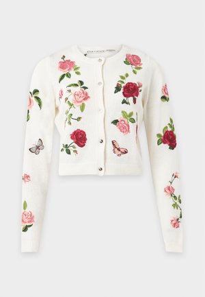 Femme aux longs cheveux foncés porte un cardigan blanc avec une broderie florale rose et rouge et un jean bleu à jambes larges avec des ourlets effilochés.