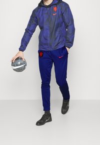 Nike Trainingsanzug mit einer dunkelblauen Jacke im Camouflage-Muster und rotem Löwenlogo, kombiniert mit blauen Hosen, die ebenfalls das Logo zeigen.
