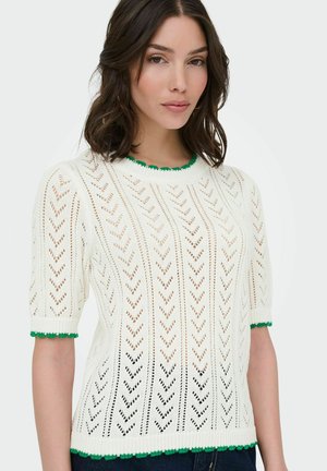 Femme aux cheveux bruns mi-longs portant un pull blanc en maille à manches courtes avec des bordures vertes au niveau de l'encolure, des poignets et de l'ourlet.