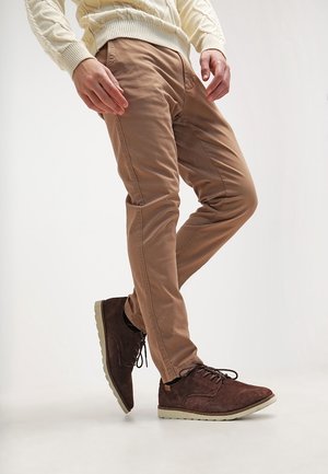 Homme portant un pantalon beige, un pull crème en maille torsadée et des chaussures décontractées en suède marron foncé avec des semelles claires, debout les jambes croisées.