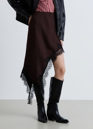 Donna che indossa una gonna marrone asimmetrica con bordo in pizzo nero, stivali neri al ginocchio, una camicia a righe e una giacca di pelle nera.
